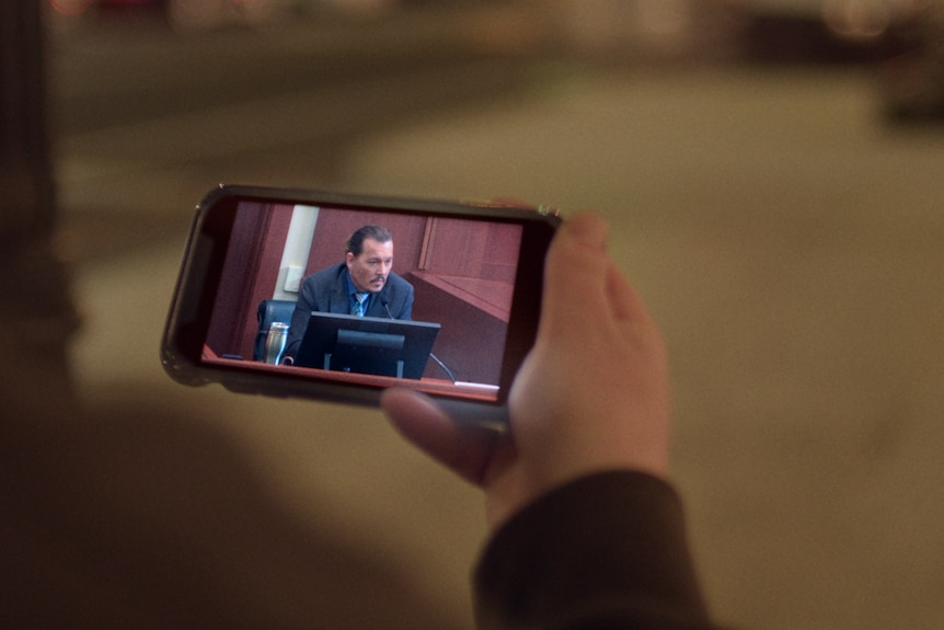 A hand holding a smartphone with Johnny Depp on the screen in court