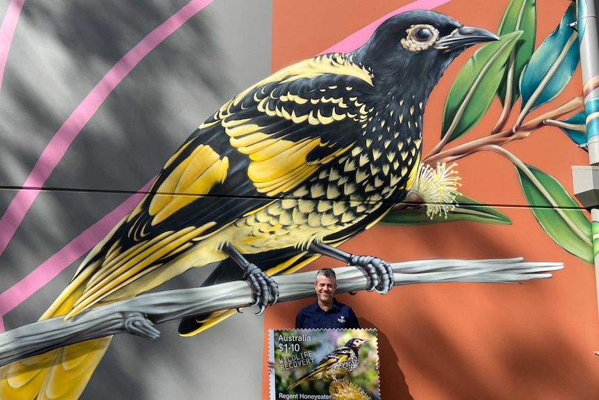 A man standing in front of a large mural of a black and yellow bird, holding a large version of a stamp.