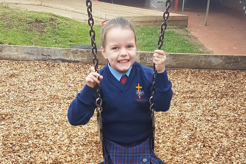 Ella sits on a swing smiling. She is wearing a school uniform and has a ribbon in her hair.