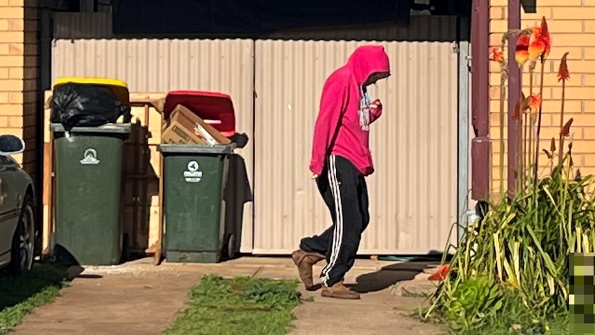 A person wearing a red hoodie and black trackpants walks past a garage with a car in the front