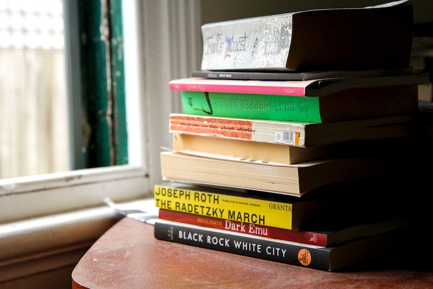 A pile of books, including Joseph Roth's The Radetzky March and A. S. Patrić's Black Rock White City, stand on a side table.