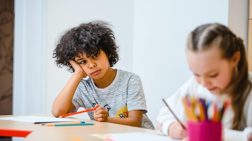 Boy leans his face on his hands looking bored while watching a girl draw.