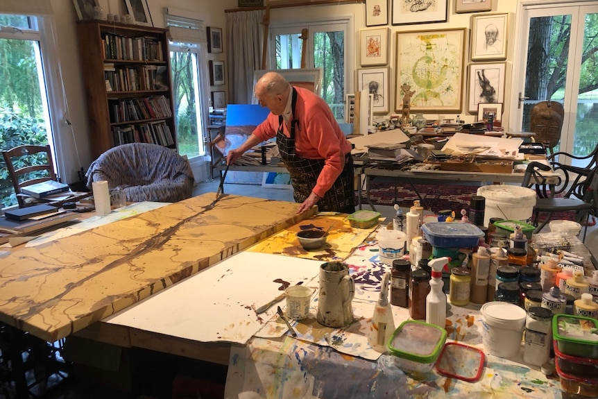 An elderly man in a pink sweater leans over an art table painting in a studio