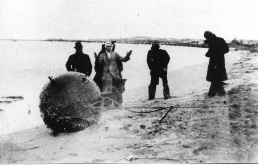 Black and white photo of men surrounding sea mine on beach