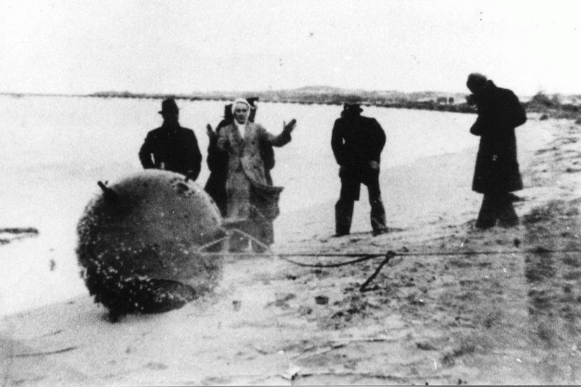 Black and white photo of men surrounding sea mine on beach.