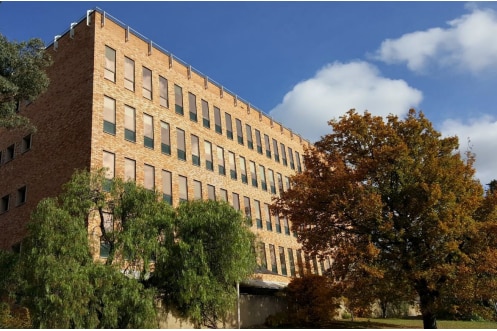 An orange brick multi-storey building surrounded by trees against blue sky and clouds.