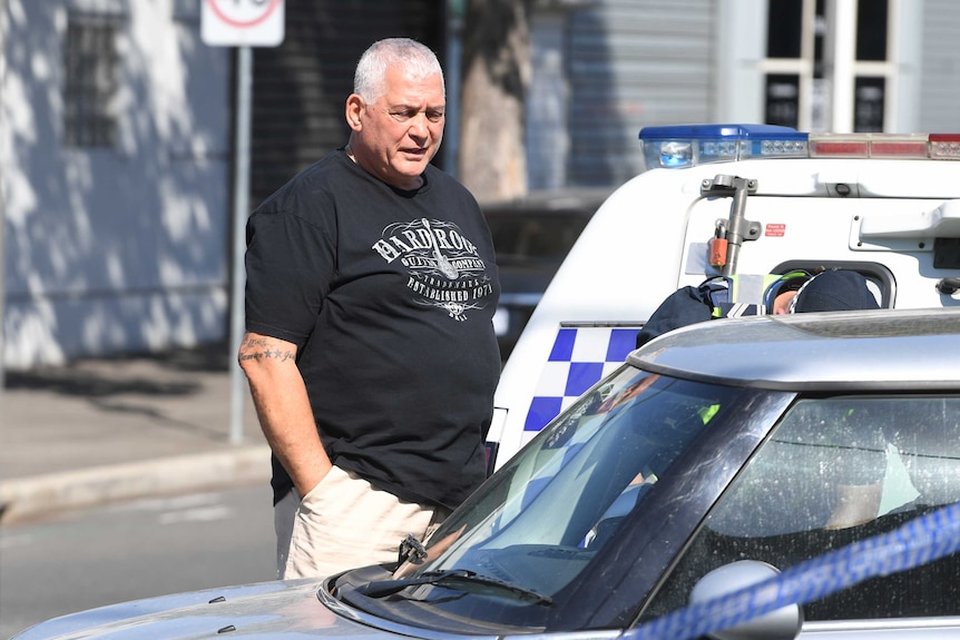 Mick Gatto, wearing black t-shirt with grey short hair standing on suburban street, with police car in background