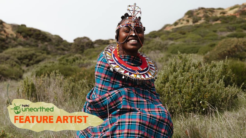 Elsy Wameyo wearing traditional clothing sits in front of a grassy cliff. She is smiling into the distance.