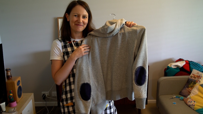 Photo of Renae wearing a black and white checked dress holding up a grey jumper that has just been mended