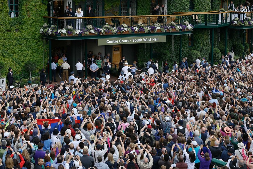 A man in a white tracksuit holds a golden trophy aloft from a balcony as people pack the area below.