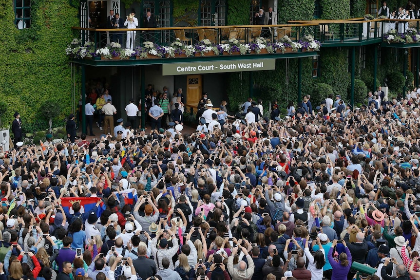 A man in a white tracksuit holds a golden trophy aloft from a balcony as people pack the area below.