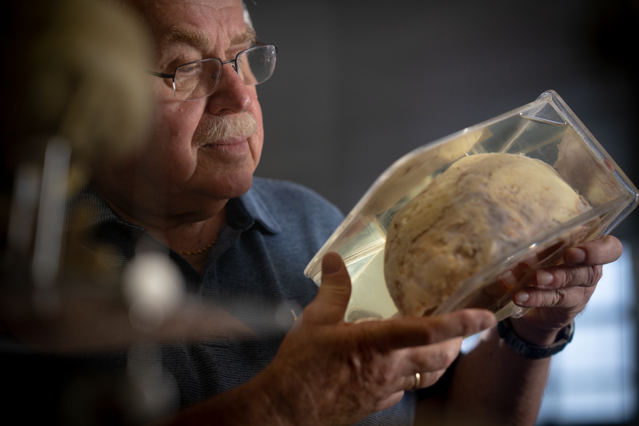 A man inside a lab studying a human skull, a skeleton model hangs nearby.