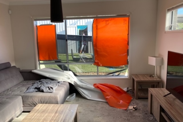 The living room of a home is covered in debris from a smashed window that is covered by an orange tarp.