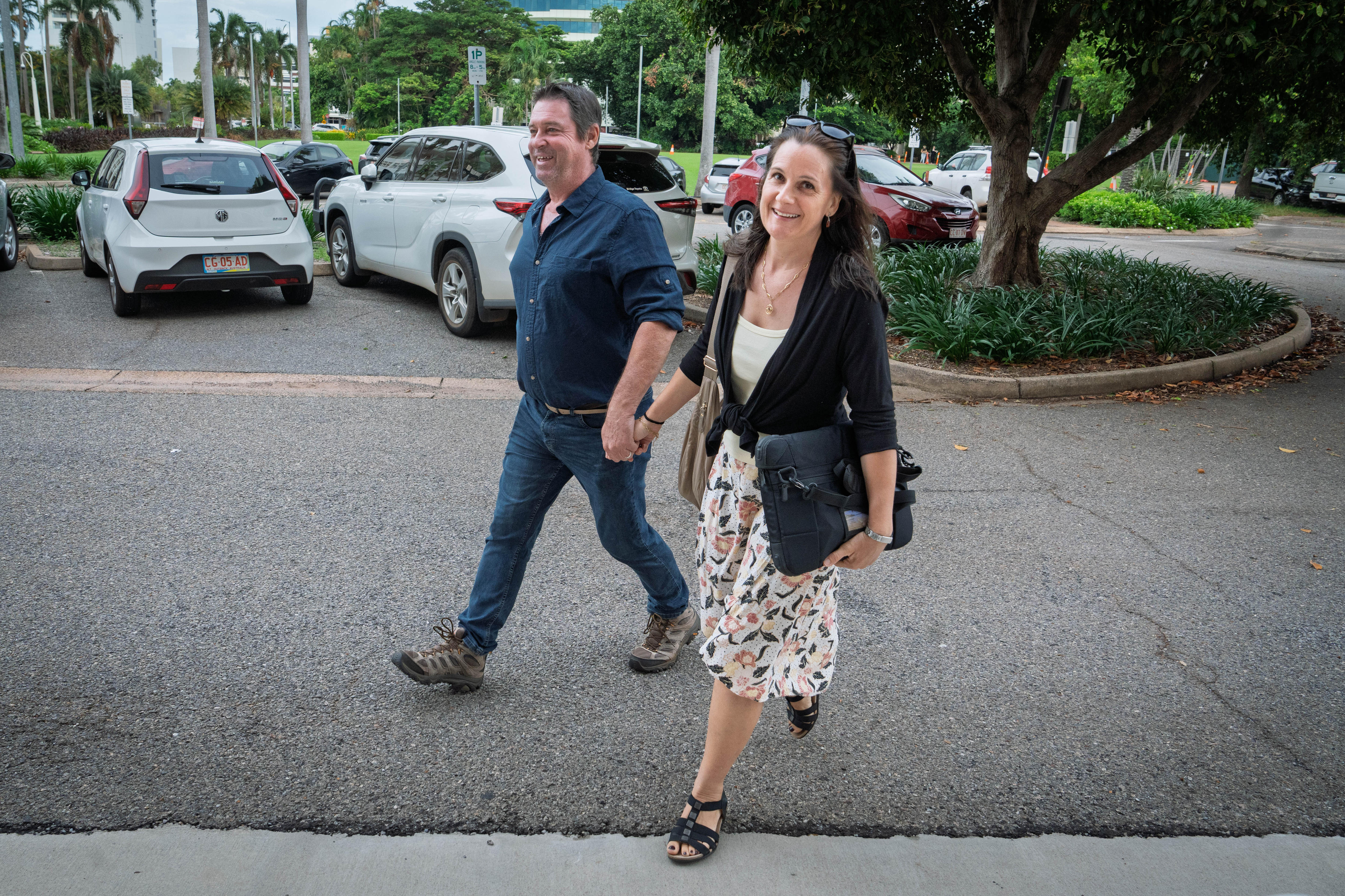 Homem branco e mulher caminhando. Homem de jeans e camisa azul escura, de mãos dadas com uma mulher de vestido branco e jaqueta preta