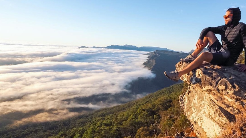 A man sits on the edge of a cliff high above clouds, or mist, covering the landscape below.