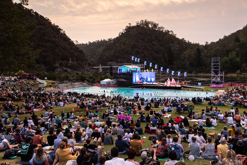 A crowd of people sitting on grass and a smaller group in a pool watching a performance on stage, forested hills in the b/g