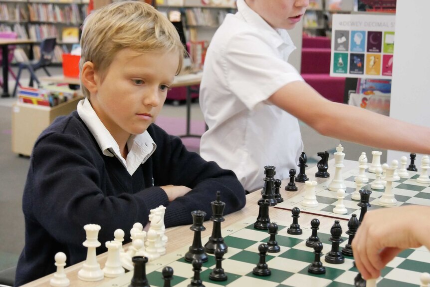 A primary school boy sits at a chess board with a look of concentration.