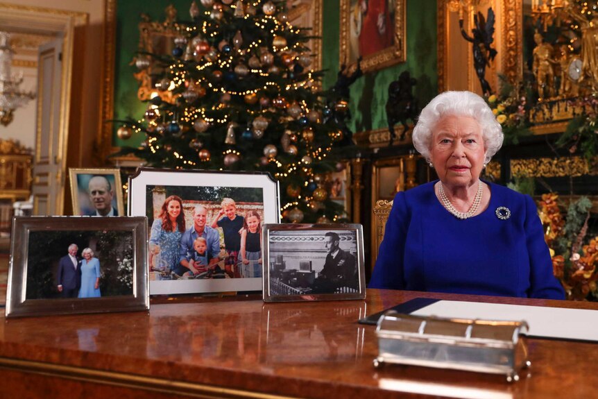 The Queen sits at a desk near a Christmas tree. Photos of Prince Charles, Prince Philip and Prince William's family are nearby.