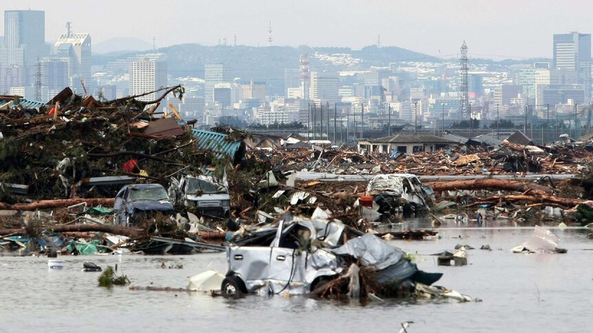 Rubble and flooded water against a city backdrop. A destroyed car floats front of frame.