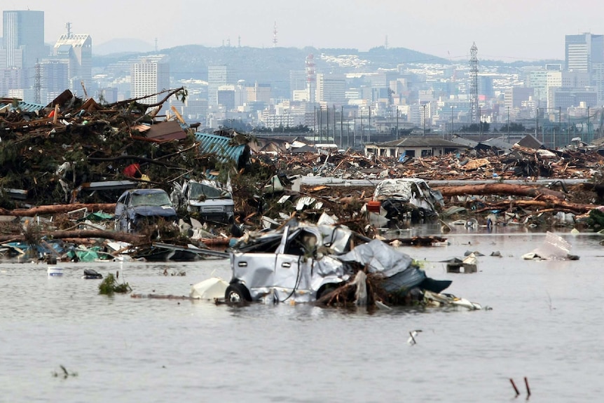 Rubble and flooded water against a city backdrop. A destroyed car floats front of frame.