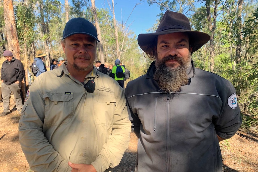 Two men wearing ranger gear standing side by side smiling. There is bushland behind them.