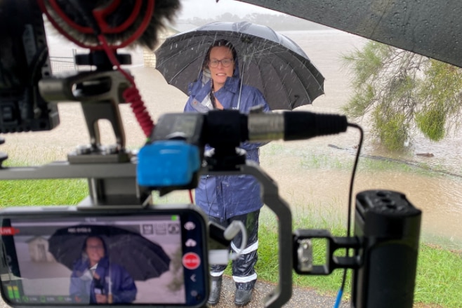 Woman standing under umbrella talking to a camera beside flooded river as heavy rain falls.