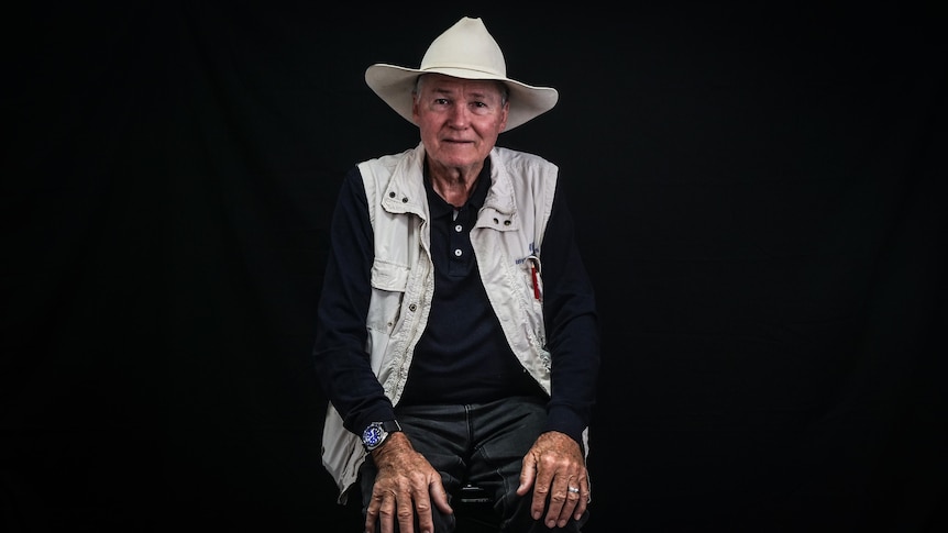 An older man in a cowboy hat and vest sitting on stool in front of a black background.