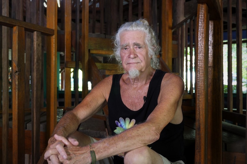 A man with white hair and white beard, black singlet, sitting on wooden steps inside a wooden building