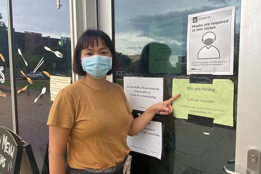 A woman wearing a blue mask and a yellow shirt standing outside a cafe and pointing to a sign which reads 'we are hiring'.