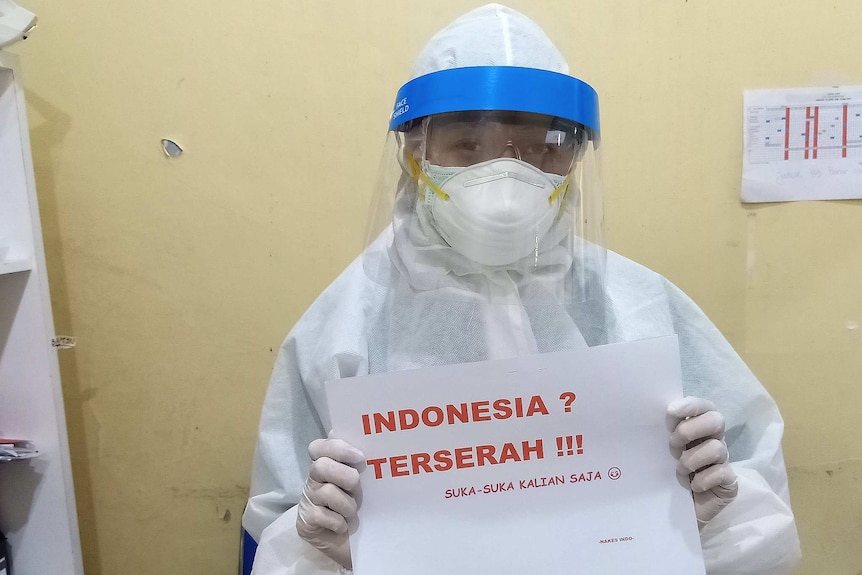 Indonesian medical worker wearing protective equipment holds sign reading "Indonesia? Whatever! Do what you like."