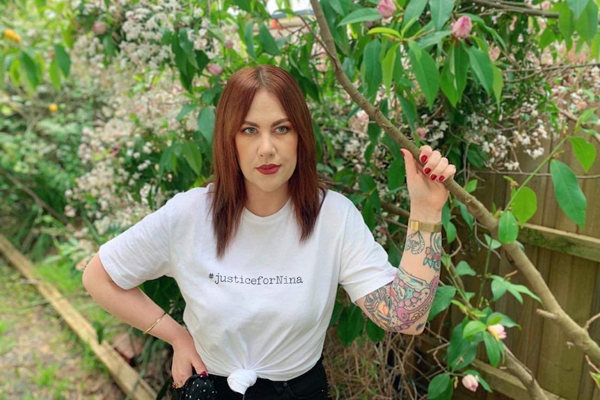 A young woman with red hair and tattoos, wearing a 'justice for Nina' t-shirt stands under a tree