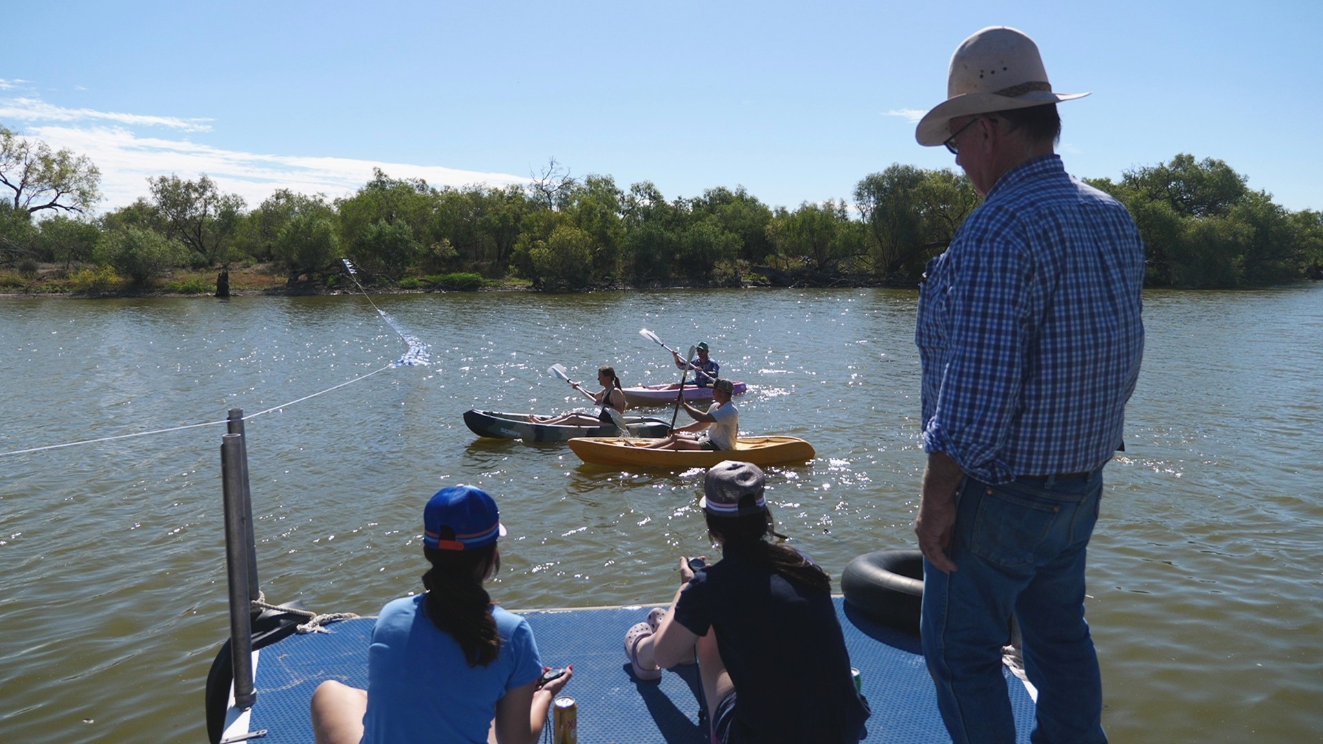 Don Raymont ao lado de duas pessoas sentadas em um cais com canoas alinhadas na água. 