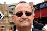 A balding Milton Orkopolous looks down the barrel of the camera, wearing oval-shaped sunglasses