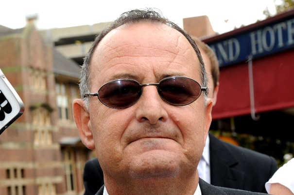 A balding Milton Orkopolous looks down the barrel of the camera, wearing oval-shaped sunglasses