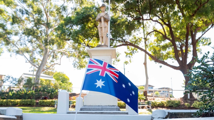 A small Australian flag stands on the ground below a tall Anzac monument