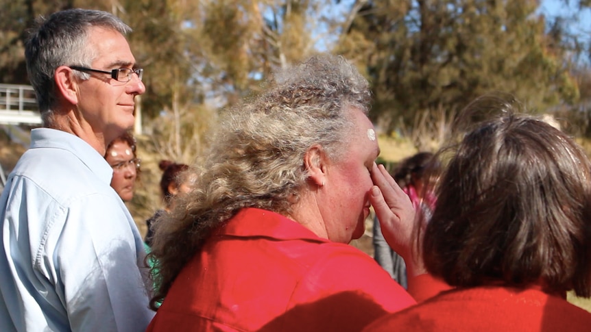 Shot a woman taken from the side, showing her crying, with paint on her forehead, other people standing in a circle