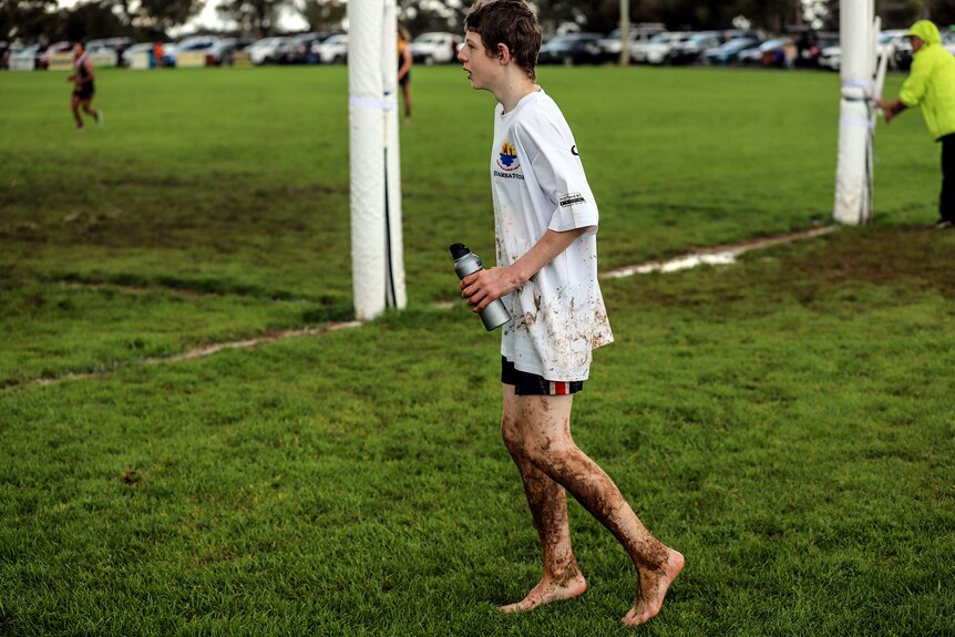 A young boy wearing white tshirt with muddy legs on a football ground
