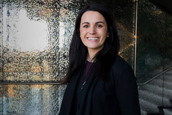 A woman stands before a cement wall wearing black jacket and black shirt, with long dark hair, smiling widely.