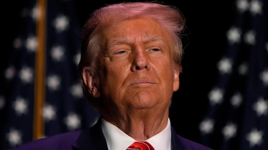 An elderly man in a blue suit, white shirt, red tie, looks on with a neutral expression