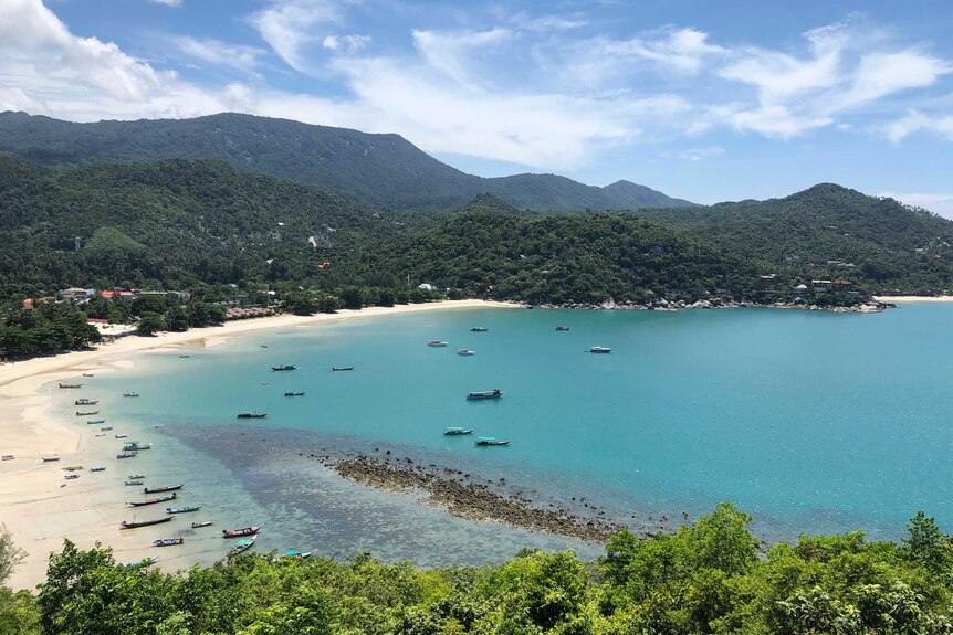 Fishing vessels and boats used to ferry tourists sit idle along a deserted beach with turquoise waters below green mountains.