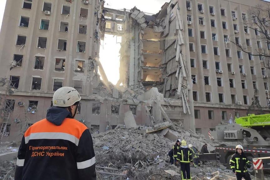 People in high visibility gear look up at a tall building that is crumbling.