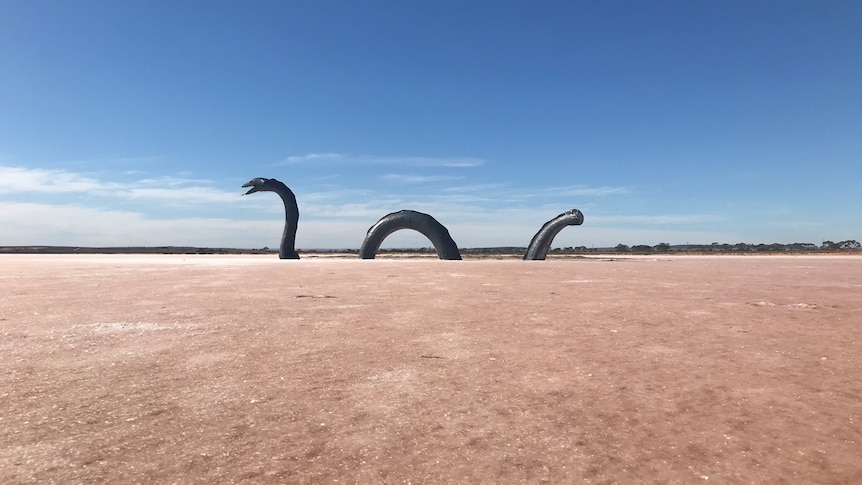 A sculpture of a large eel stands side-on, embedded in a pale pink salt lake base. The sky is blue with some clouds. 