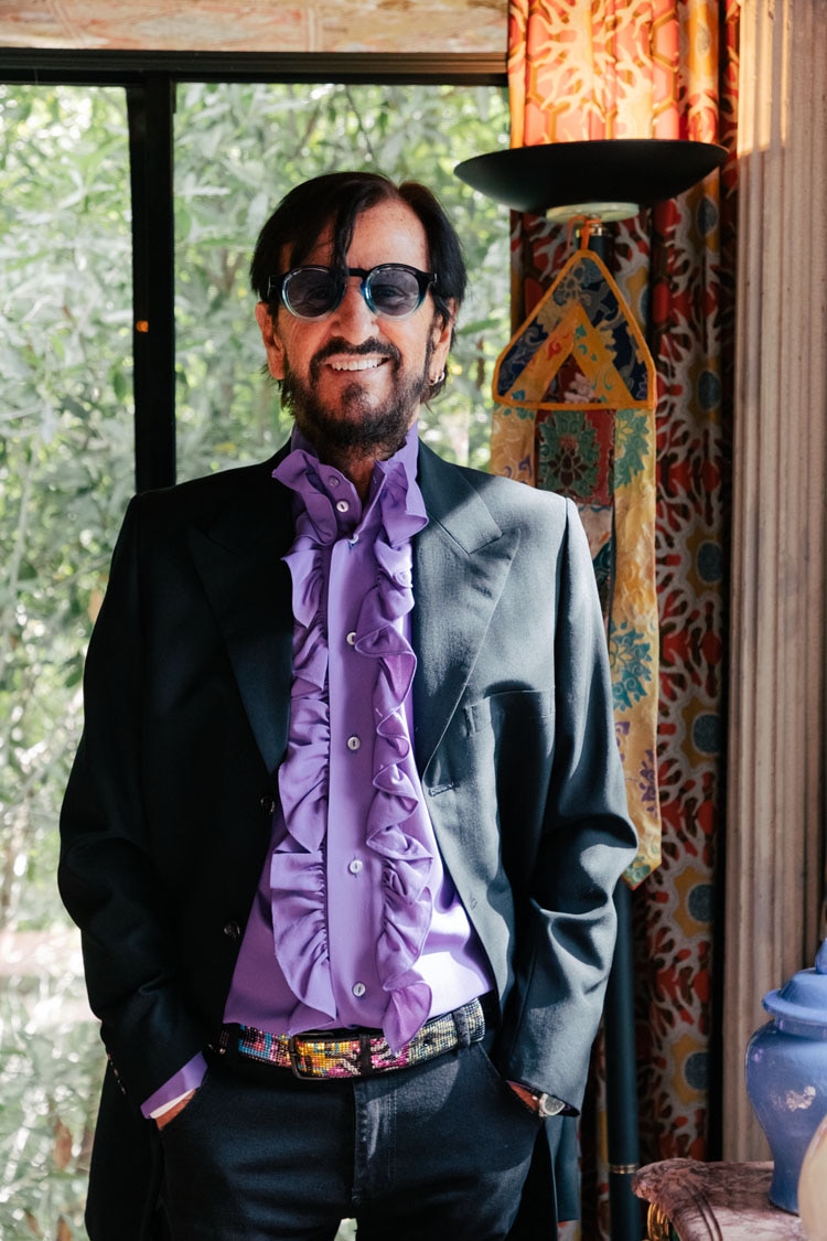 Ringo Starr se levanta, sonriendo y con las manos en los bolsillos. Lleva una camisa morada, una chaqueta oscura y gafas de sol.