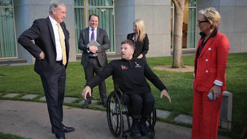 Richard Colbeck stands with his hands on his hips, looking down at Dylan Alcott who has his hands stretched oueither side of him