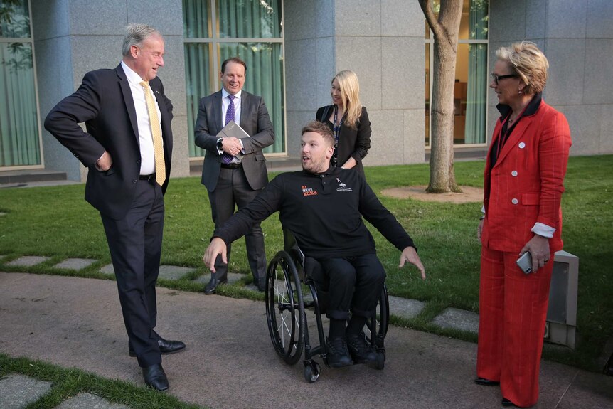 Richard Colbeck stands with his hands on his hips, looking down at Dylan Alcott who has his hands stretched oueither side of him