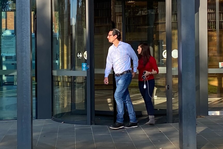 A man in a business shirt and a woman in a red top leave through a revolving door. 