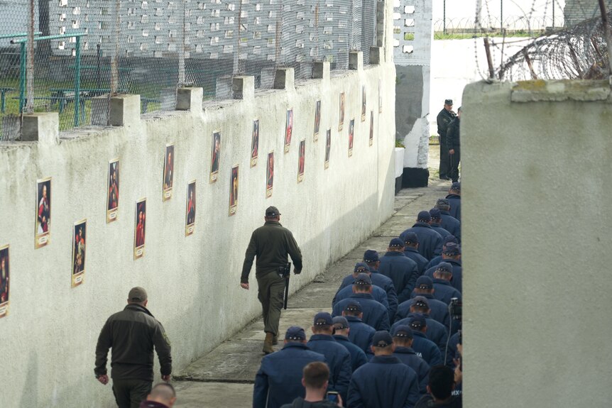 Охранники идут рядом с заключенными, сидящими в бетонном переулке внутри украинского изолятора для военнопленных.