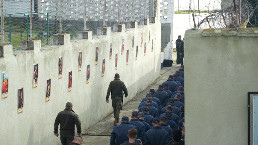 Guards walk alongside seated prisoners in a concrete alleyway inside a Ukrainian prisoner of war facility.