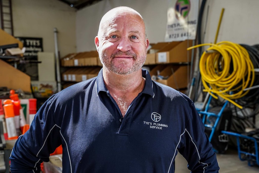 A man wearing a navy polo shirt in a large shed with plumbing gear behind him smiles for a photo.