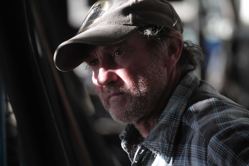 Close up of Peter Clarke standing in the dairy farmer wearing a battered cap and blue-grey flannel shirt, looking off camera